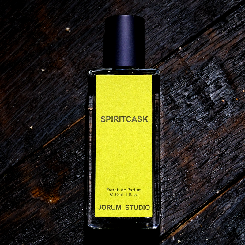 Perfume bottle, sitting on a charred wooden floor, with green label with the fragrance name "Spiritcask" written across the middle of the label.