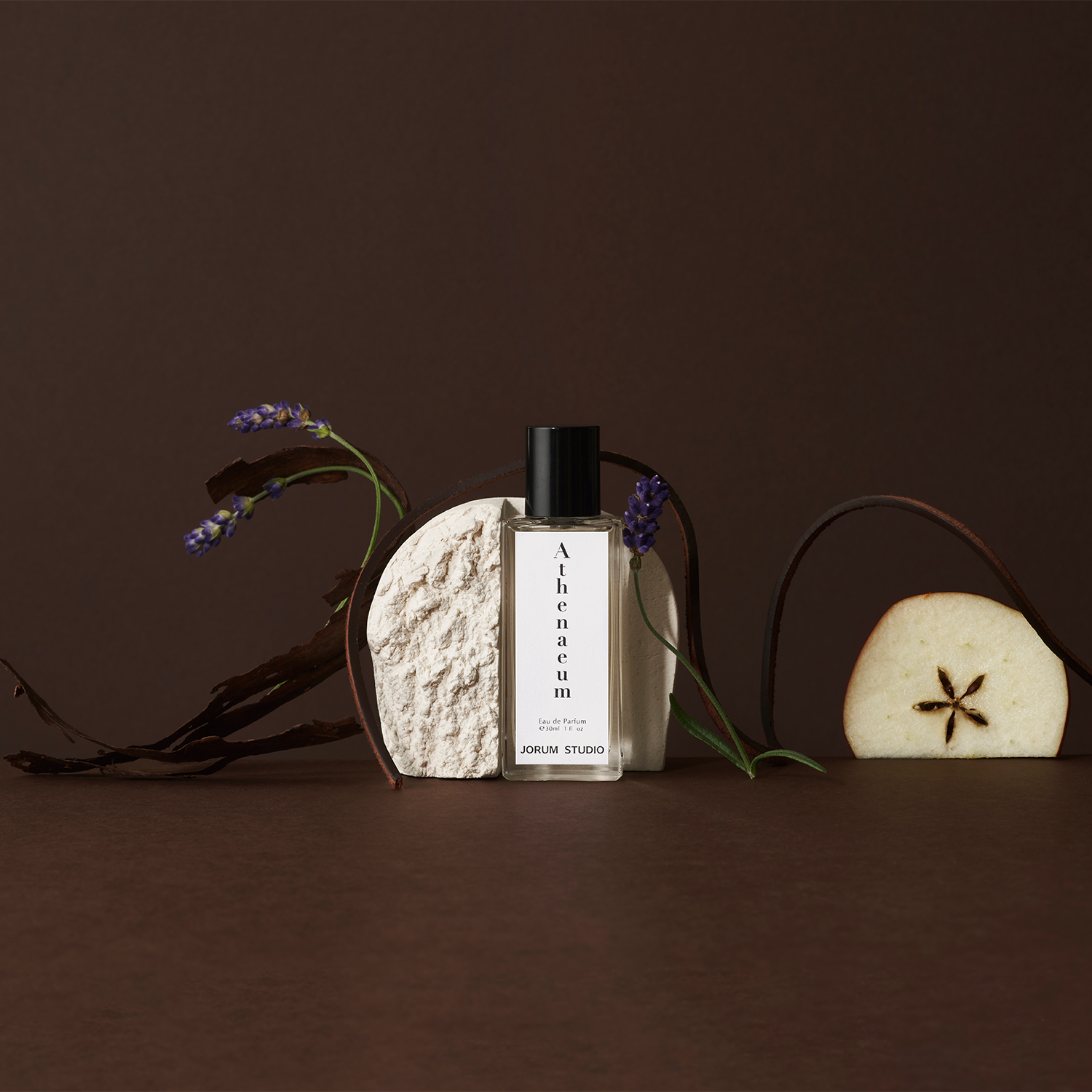 Jorum Studio Athenaeum perfume bottle arranged in a still-life with white stone, apple slice, lavender and wood shavings against a brown background