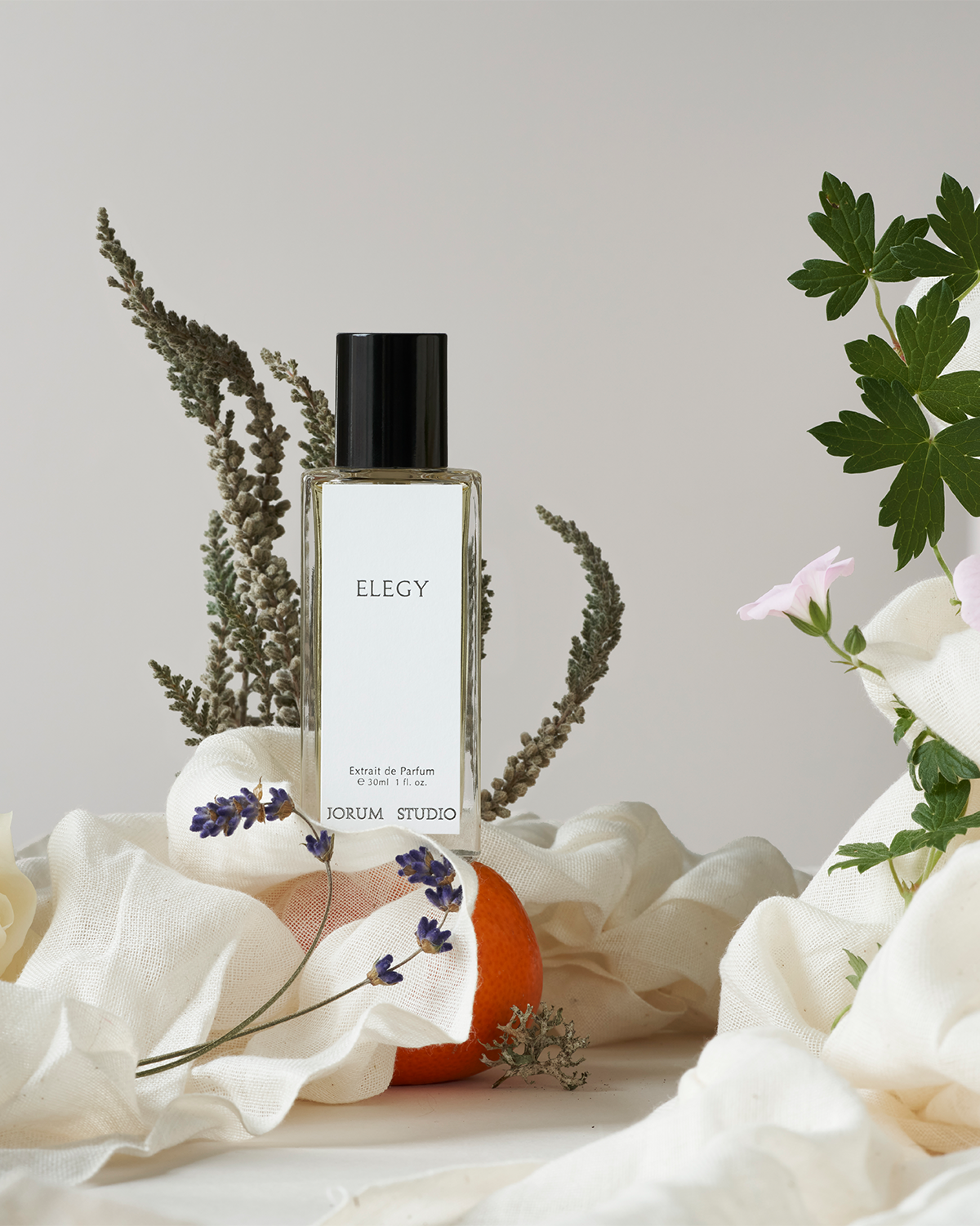 Jorum Studio Elegy perfume bottle arranged in a still-life featuring white fabric, lavender and heather flowers and an orange.