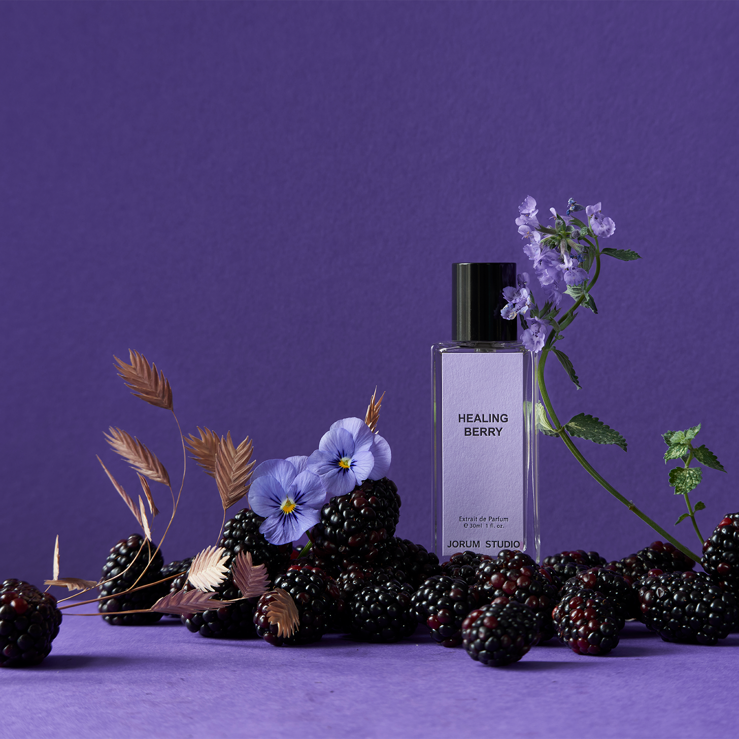 Jorum Studio Healing Berry perfume bottle arranged in a still-life featuring blackberries, violets and barley grains against a purple background