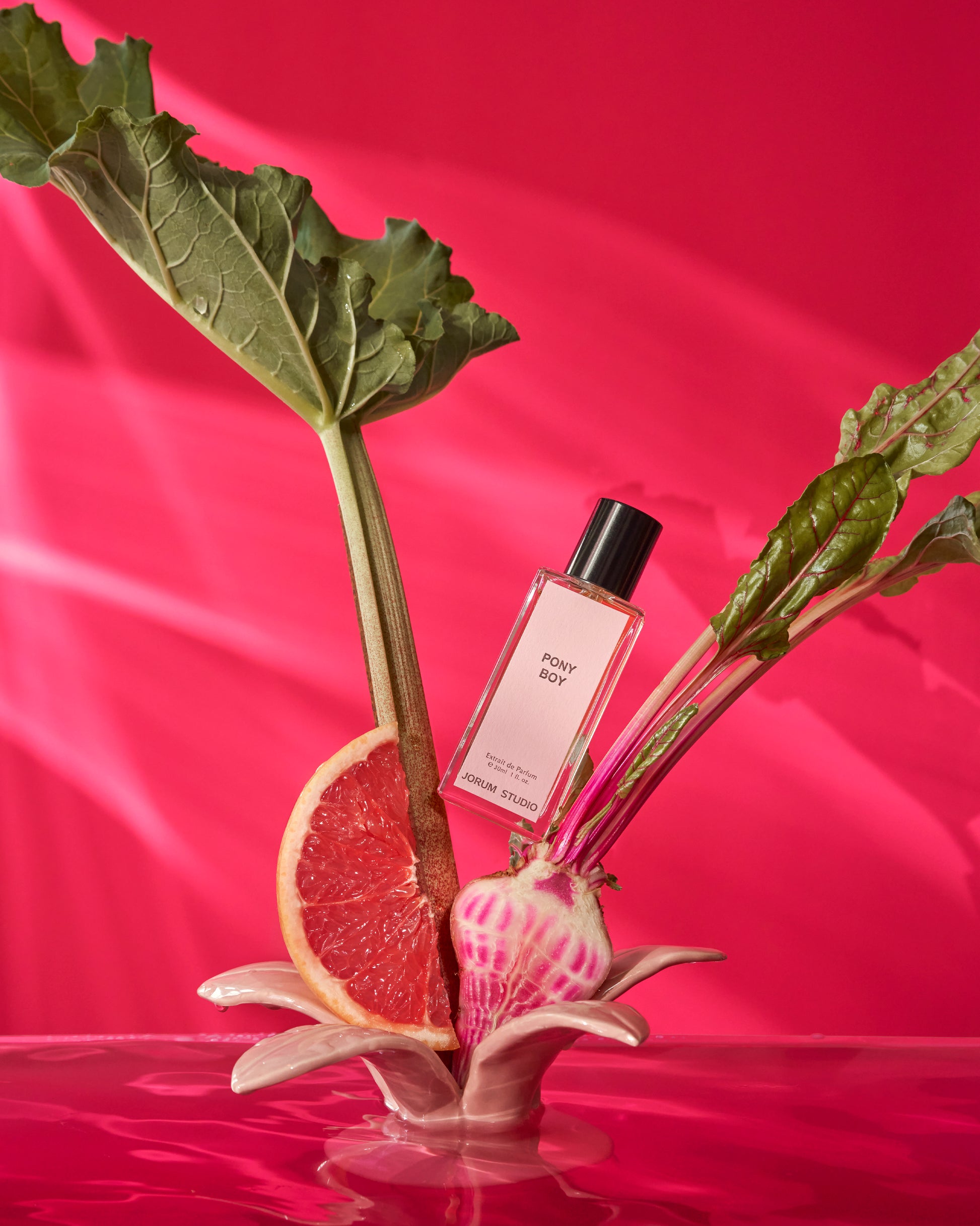 Jorum Studio Pony Boy perfume bottle arranged in a still-life featuring rhubarb stalks, a grapefruit wedge, sliced beetroot against a watery hot pink background