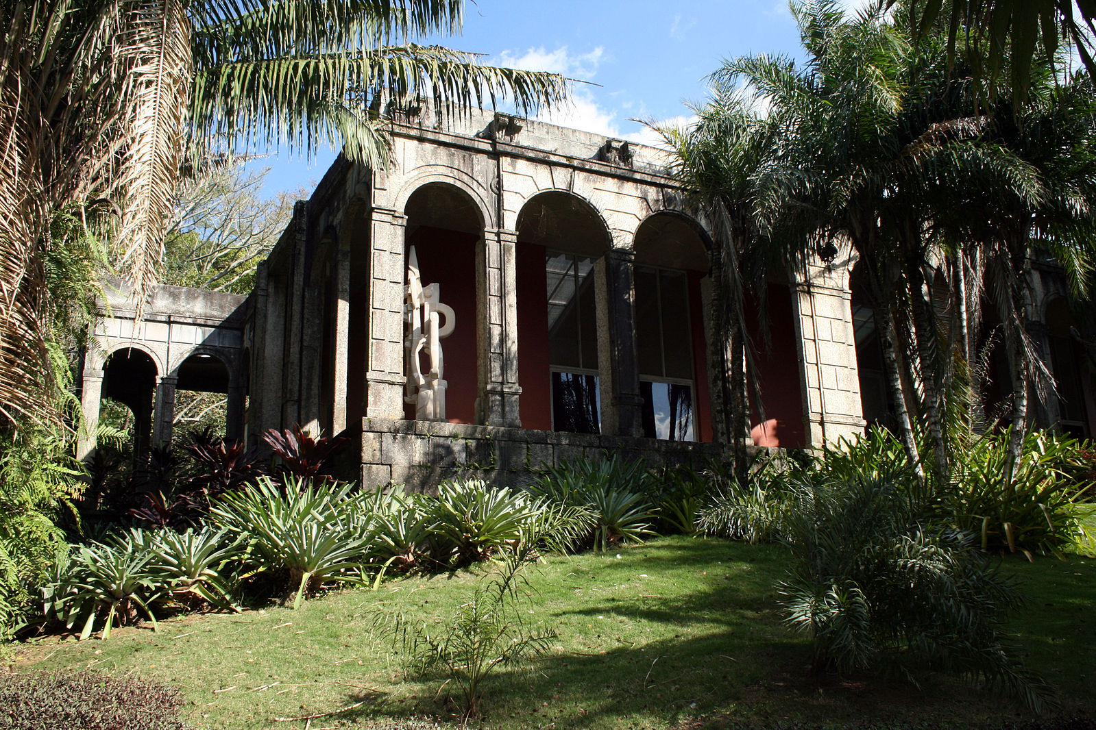 A large stone building with arches surrounded by lush greenery and palm trees