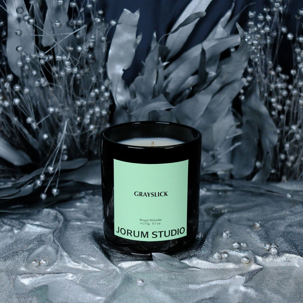 Jorum Studio Grayslick candle arranged in front of silver foliage on silver fabric made to look like frozen water