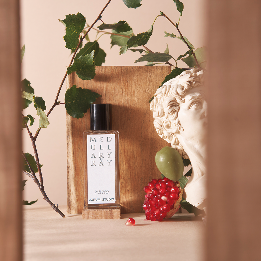 Jorum Studio Medullary-Ray perfume bottle arranged in a still-life featuring a marble bust, wooden block, olives, pomegranate seeds and fig leaves