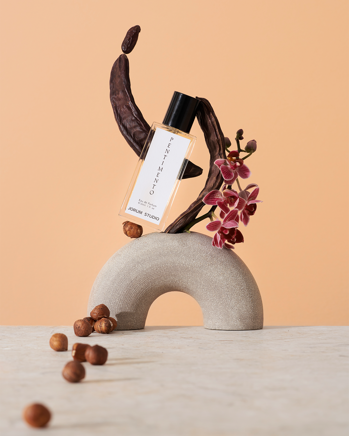 Jorum Studio Pentimento perfume bottle arranged in a still-life featuring carob pods, orchids and hazelnuts balanced on a stone semicircle